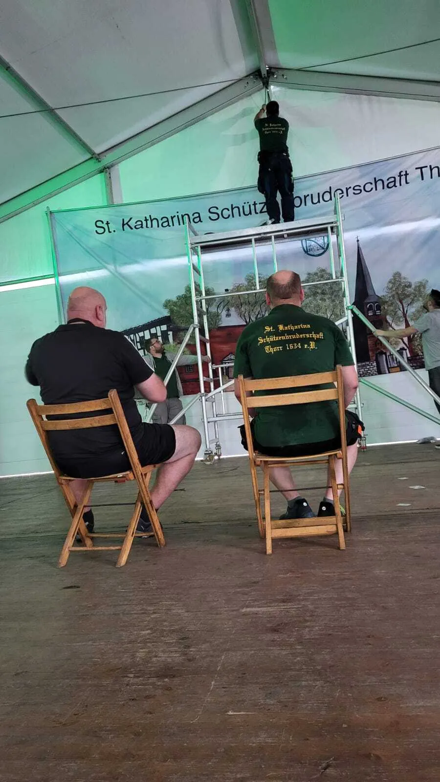 Zwei Schützenbrüder schauen zu, wie das Vereinsbanner „St. Katharina Schützenbruderschaft Thorr" hinter der Bühne angebracht wird