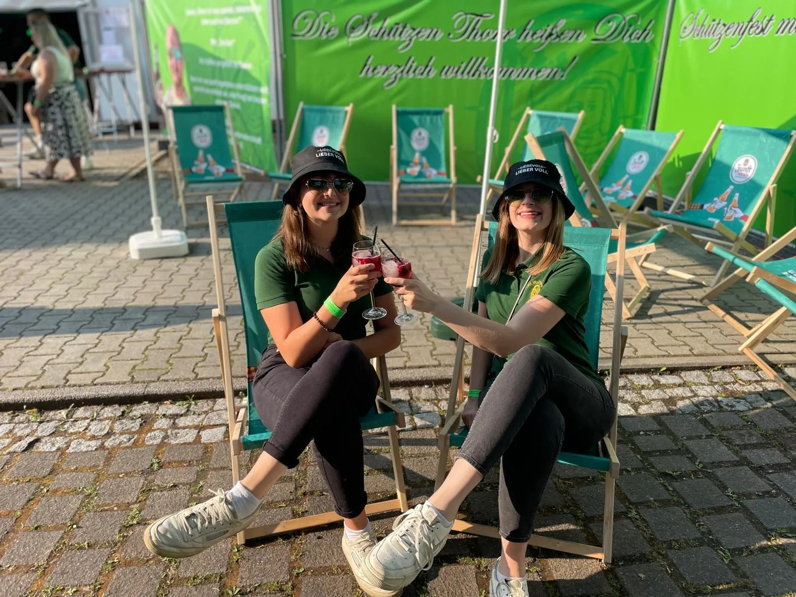 Zwei Schützenschwestern stoßen mit Cocktails in Liegestühlen vor dem Willkommensbanner am Festzelt an