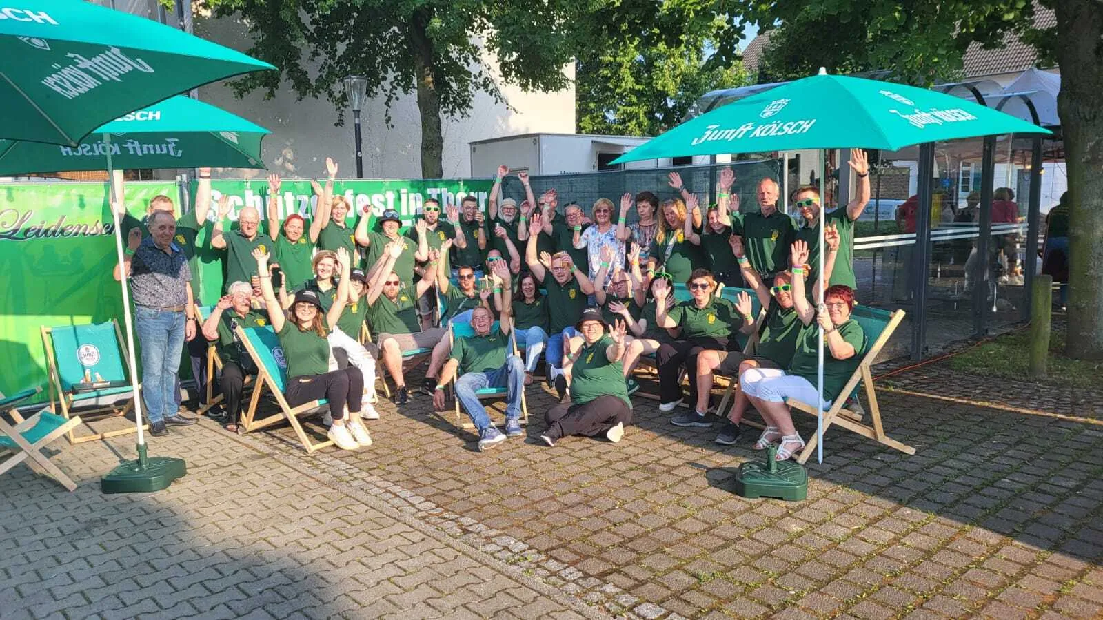 Gruppenbild der gesamten Bruderschaft beim Schützenfest 2024 vor Junft-Kölsch-Schirmen und Liegestühlen, viele Hände gehen jubelnd nach oben