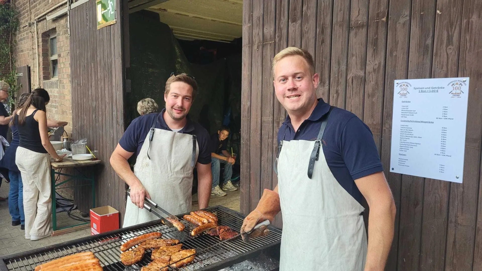 Zwei Schützenbrüder mit weißen Schürzen am Holzkohlegrill mit Steaks und Würstchen, daneben eine Speisen- und Getränkekarte zum 100-jährigen Jubiläum einer befreundeten Bruderschaft