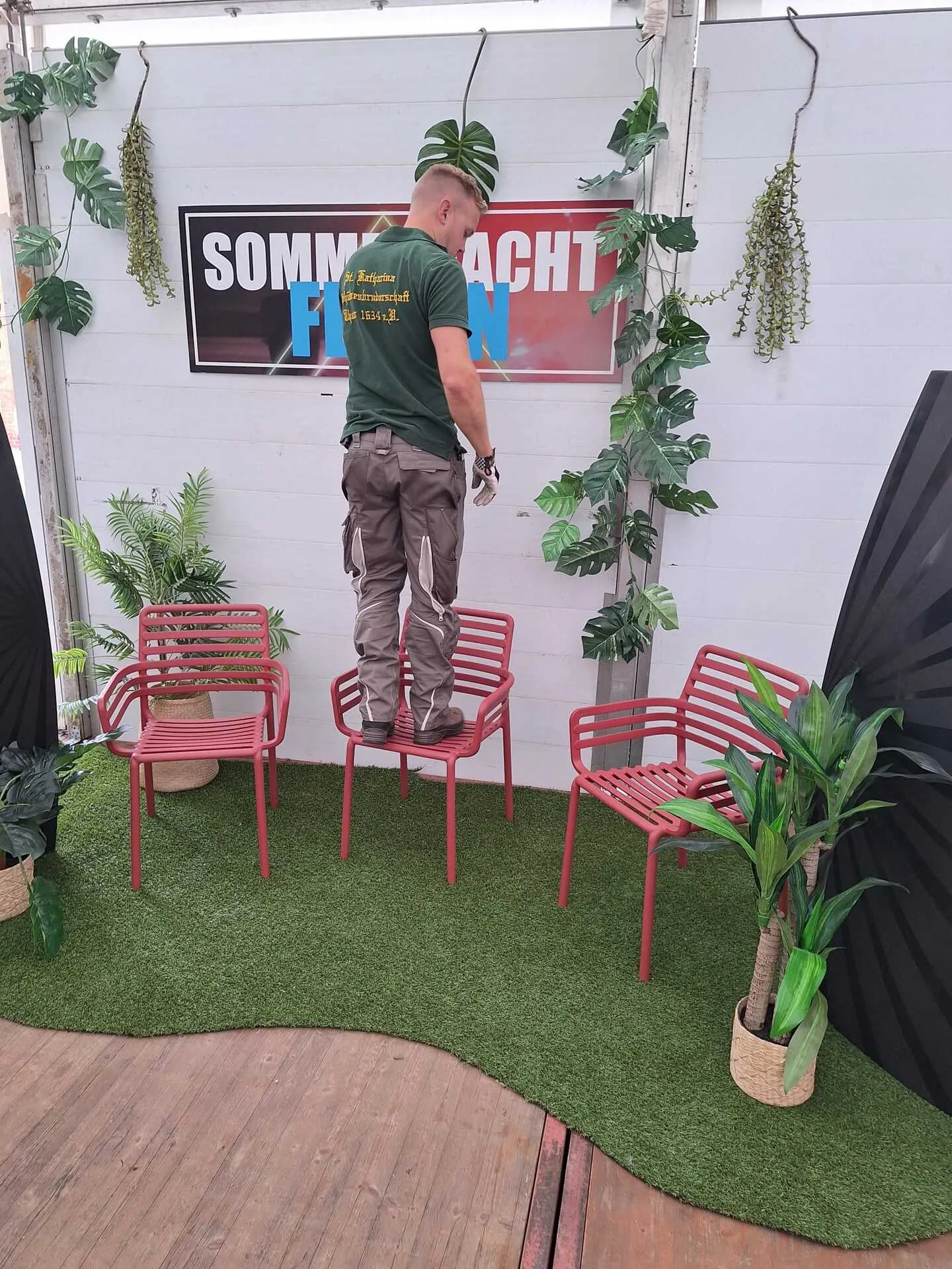 Schützenbruder hängt das „SOMMERNACHT FEIERN"-Banner über drei roten Stühlen mit Kunstrasen-Foto-Set für die Sommernacht 2025 auf