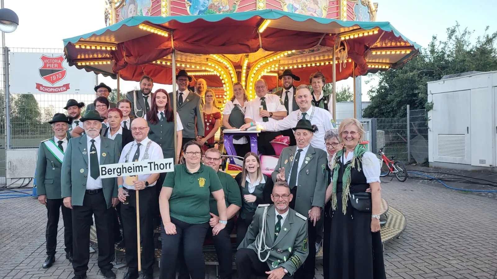 Gruppenbild der Bruderschaft mit Schild „Bergheim-Thorr" vor einem Karussell beim Bezirks-Schützenfest in Schophoven